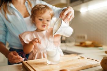 Young mother and kid pours milk into a glass, ingredients for pastry. Woman and little girl cooking on the kitchen, cake preparation. Happy family makes sweet dessert at the counter