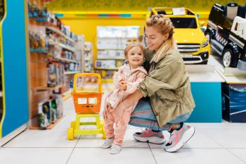 Happy mother with little daughter in kids store. Mom and child together choosing toys in supermarket, family shopping