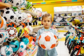 Little boy with ball in kids store, front view. Son choosing toys in supermarket, family shopping, young customer