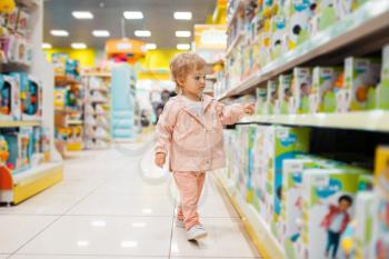 Little girl at the shelf choosing toys in kids store, side view. Daughter in supermarket, family shopping, young customer