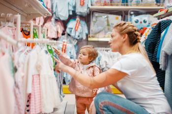 Mother with her little girl choosing clothes in kids store. Mom and child buying dress in supermarket together