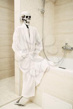 Human skeleton in white bathrobe sitting on the edge of the bath in bathroom, black humor