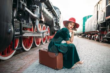 Woman in red hat sitting on suitcase against vintage steam train. Old locomotive. Railway engine, ancient railroad vehicle