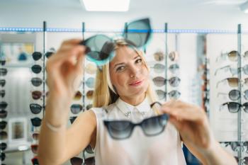 Female buyer chooses sunglasses in optics store, showcase with spectacles on background. Selection of eyes protection in glasses shop, optometry concept