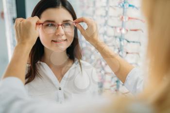 Female optician and customer chooses glasses frame against showcase with eyeglasses in optics store. Selection of spectacles with professional optometrist