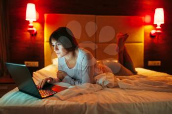 Happy woman lying in bed and uses laptop, good morning, bedroom interoir on background. Lady waking up
