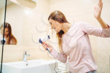 Young woman pampering with hairdryer at the mirror in bathroom. Female person makes hairstyle in the morning.