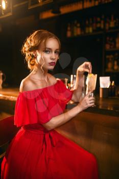 Beautiful woman in red dress drinks alcohol cocktail at the bar counter. Date in nightclub, attractive love couple in pub