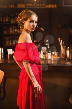 Portrait of young woman in red dress standing at the bar counter. Beautiful lady with beverage in hand in club