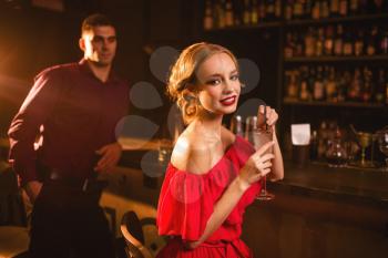 Smiling woman in red dress with cocktail in hand, man behind bar counter, flirting. Date in nightclub, attractive love couple in pub