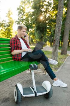 Young man with gyroboard sitting on the bench in park. Outdoor recreation with electric gyro board. Eco transport with balance technology, electrical gyroscope vehicle