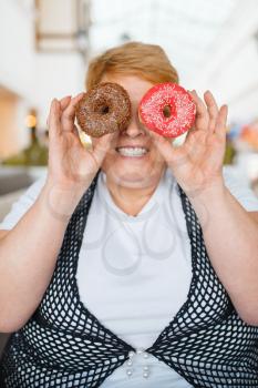 Fat woman holding doughnuts instead of eyes in mall restaurant, unhealthy food. Overweight female person at the table with junk dinner, obesity problem