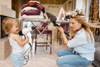 Housewife with little kid playing vacuum cleaner and hair dryer at the ironing board. Woman with child doing housework at home together. Female person with daughter having fun in their house