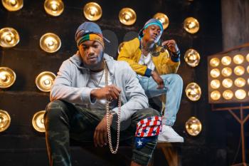 Two black rappers sitting on the steps, perfomance on stage with spotlights on background. Rap performers on scene with lights, underground music, urban style