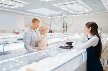 Happy love couple buying gold decoration. Male and female consumers looking on jewels in jewelry store. Man and woman choosing wedding rings. Future bride and groom in jewellery shop