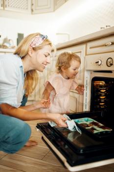 Young mother and kid remove from the oven baking sheet with chocolate pastry. Woman and little girl cooking on the kitchen, cake preparation. Happy family makes a dessert