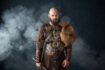Bearded viking with axe dressed in traditional nordic clothes, barbarian image. Ancient warrior in smoke on dark background