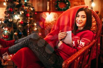 Young woman sitting on a chair near fireplace, christmas tree with decoration on background, xmas holiday celebration