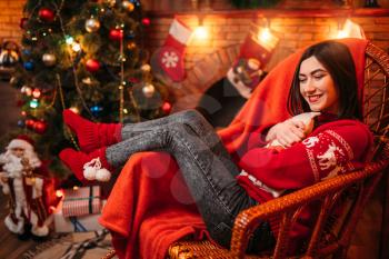 Young woman sitting on a chair near fireplace, christmas tree with decoration on background, xmas holiday celebration 