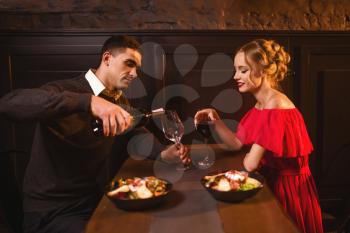 Man pours wine into a glass, young love couple in restaurant, romantic date. Elegant woman in red dress and her man, anniversary celebration