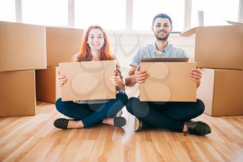 Happy couple with cardboard boxes in hands sitting on the floor, moving to new home, housewarming