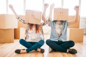 Playful couple with cardboard boxes on their heads sitting on the floor, moving to new home, housewarming. Man and woman have a fun