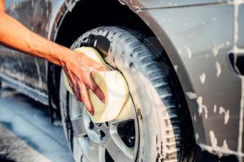 Male hand rubbing car wheel with foam, automobile in suds. Carwash station