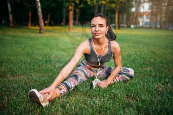 Girl in sportswear and headphones sitting on grass in summer park. Sporty girl on outdoors morning workout