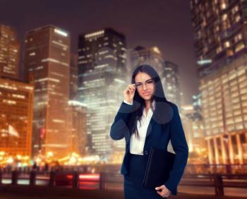 Portrait of young, beautiful businesswoman in glasses and suit with laptop in hands, night cityscape on background. Modern building, financial center. Successful female businessperson
