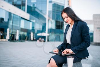 Business woman works on laptop outdoor. Modern building, financial center, cityscape. Female businessperson in suit