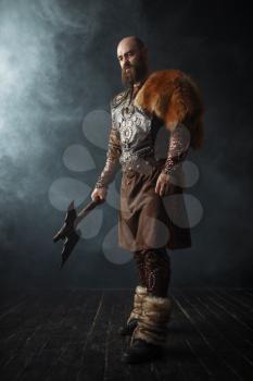Handsome viking with axe dressed in traditional clothes, nordic barbarian image. Ancient warrior in smoke on dark background