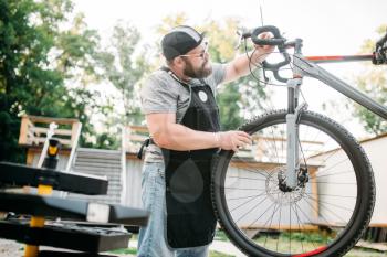 Professional bicycle mechanic in apron adjusts bike. Cycle workshop outdoor. Bicycling sport, bearded service man work with wheel