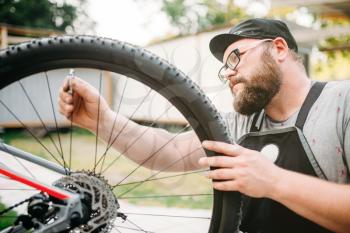 Bicycle mechanic in apron adjusts bike spokes with service tools. Cycle workshop outdoor. Bicycling sport, bearded repairman