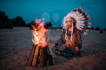Young American Indian woman against fire, Cherokee, Navajo. Headdress made of feathers of wild birds. Night ritual