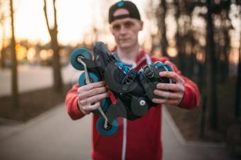 Male person shows roller skates, city park on background. Male rollerskater with rollerskates