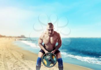 Muscular male athlete exercise with weight on sandy beach, blue sky and sea on background. Fitness training with weight. Bodybuilding workout