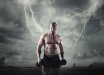 Muscular male athlete with dumbbells, lightning in dark sky on background. Fitness training with weight. Bodybuilding workout