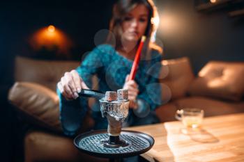 Young woman holds coal with tongs, smoking hookah at the bar, tobacco smokes at night club