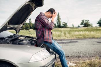 Depressed man sitting on a hood of broken car. Vehicle with open hood on roadside