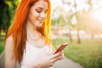 Smiling young woman with mobile phone outdoor in park on sunset. Happy girl with beautiful red hair in summer sunny day