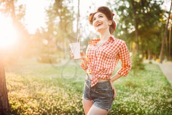 Pin up girl holds cardboard cup with a straw, city park on background. Vintage american fashion. Attractive woman in pinup style