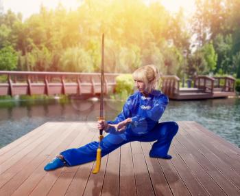 Female wushu fighter with blade against lake and green nature, martial arts. Woman in blue cloth on outdoor fight training