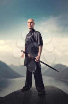 Wushu master with sword poses on the top of mountain, martial arts. Man in black  cloth poses with blade