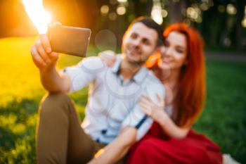 Love couple makes selfie on phone camera in summer park on sunset. Romantic date of attractive woman and young man