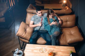 Young couple smoking hookah on leather couch at the bar, tobacco smoke and relaxation
