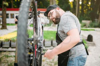 Repairman works with bike wheel, cycle workshop outdoor. Bearded bicycle mechanic in apron
