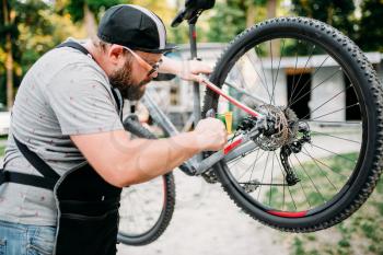 Bicycle mechanic in apron adjusts with service tools back disk brakes. Cycle workshop outdoor. Bicycling sport, bearded repairman