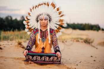 American Indian woman sitting in yoga pose. Headdress made of feathers of wild birds. Cherokee, Navajo culture, ethnic peoples traditions