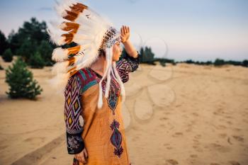 Beautiful American Indian girl in native costume looks in a distance outdoors. Headdress made of feathers of wild birds. Cherokee, Navajo culture, ethnic peoples