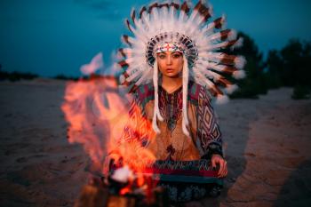 American Indian girl against bonfire in the night, female shaman, Cherokee, Navajo. Headdress made of feathers of wild birds. Traditional ritual